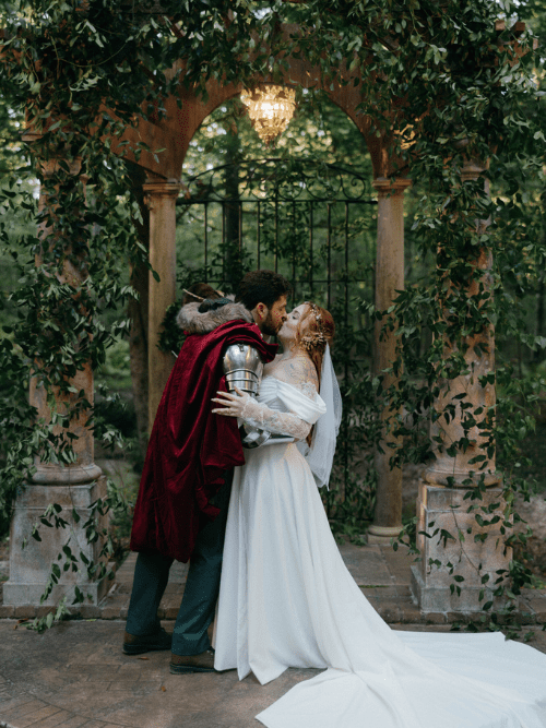 Woodland Wedding Central Arkansas A couple dressed as a medieval knight and a bride kiss under a vine-covered stone archway in a garden, surrounded by greenery and soft natural light.