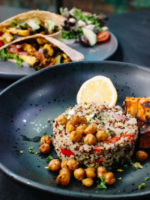 Mediterranean Cuisine to Arkansas Home Cooks A black plate with quinoa salad topped with roasted chickpeas, a lemon wedge, and roasted sweet potatoes. In the background, another plate holds tacos with vegetables and a side salad.