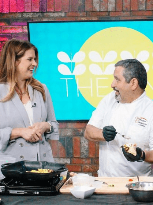 Mediterranean Cuisine to Arkansas A woman in a light gray blazer smiles and talks with a chef in a white uniform and black gloves, who is preparing food at a counter on a cooking show set with a teal and yellow sign in the background.