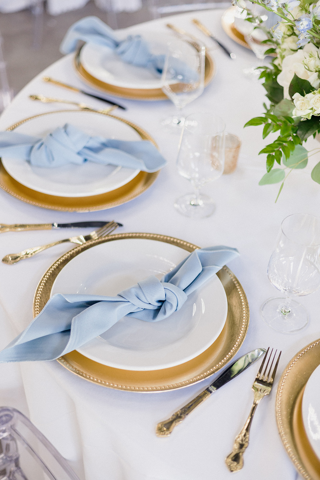 Catering Menu Near Me Elegant table setting with gold chargers, white plates, light blue knotted napkins, silver cutlery, clear glasses, and a floral centerpiece on a white tablecloth.