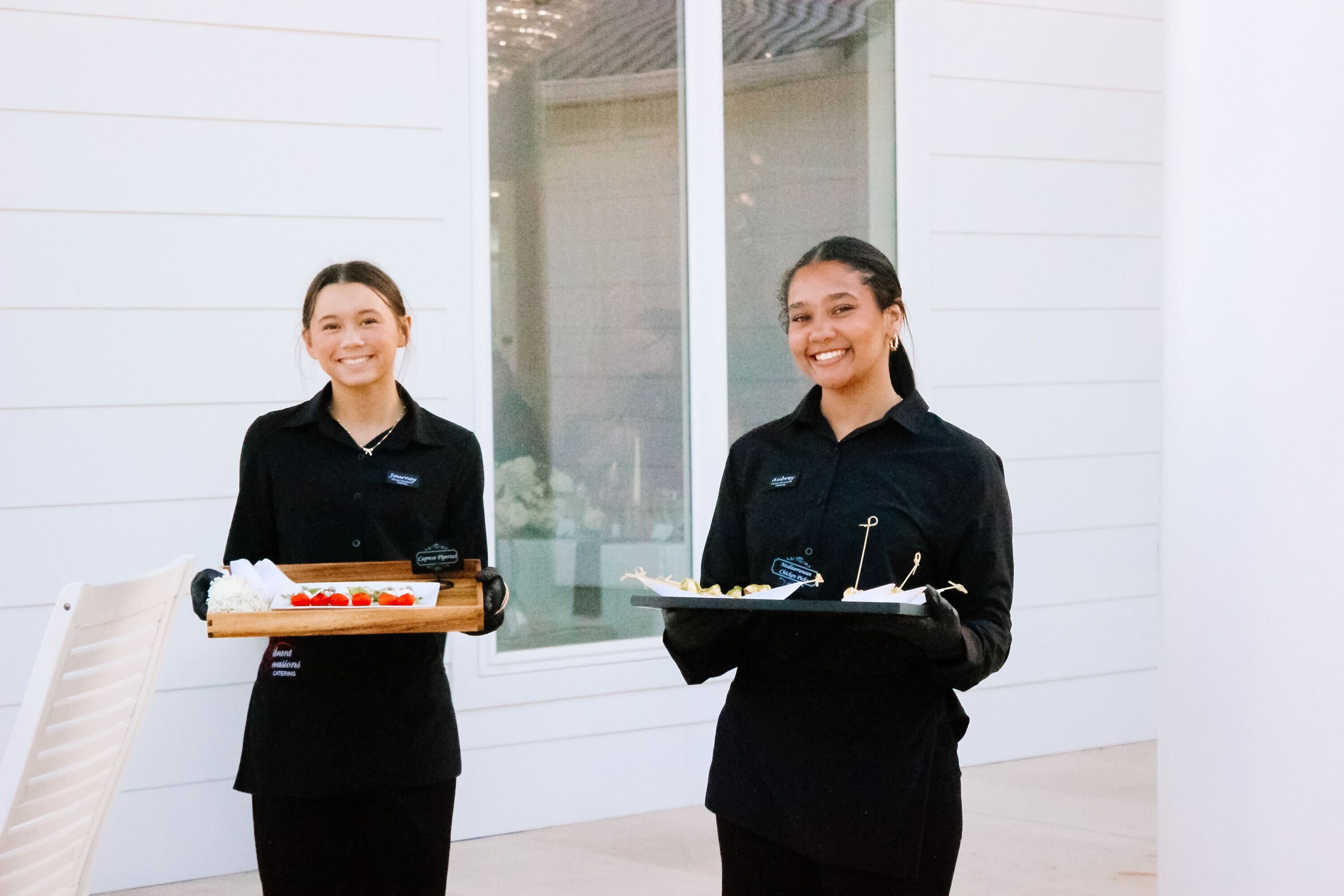 Two smiling servers in black uniforms hold trays of appetizers outdoors in front of a white building with large windows at Legacy Acres in Conway, Arkansas.