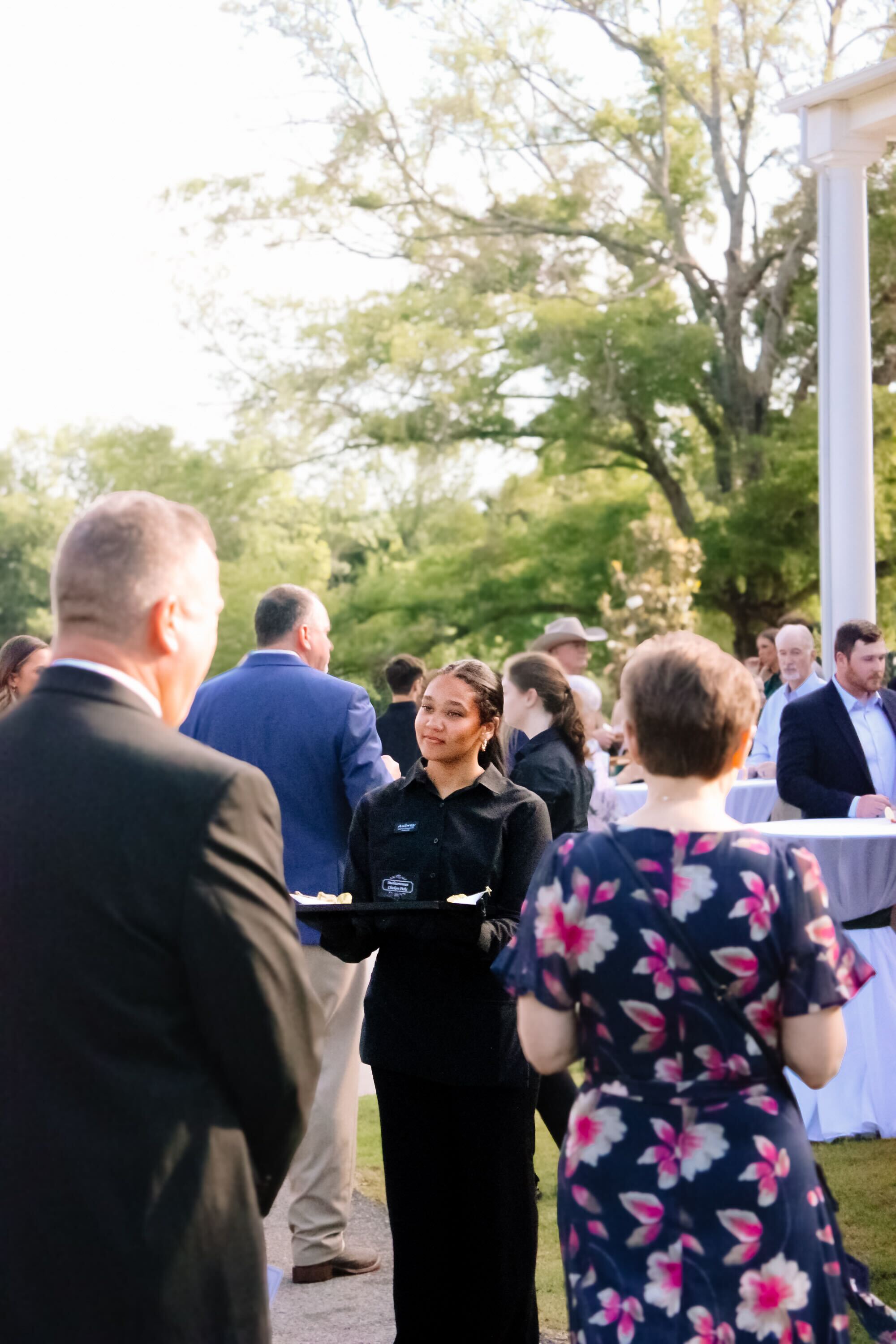 A server in black holds a tray of food at an outdoor event, while guests in formal and floral attire mingle under trees and sunlight.