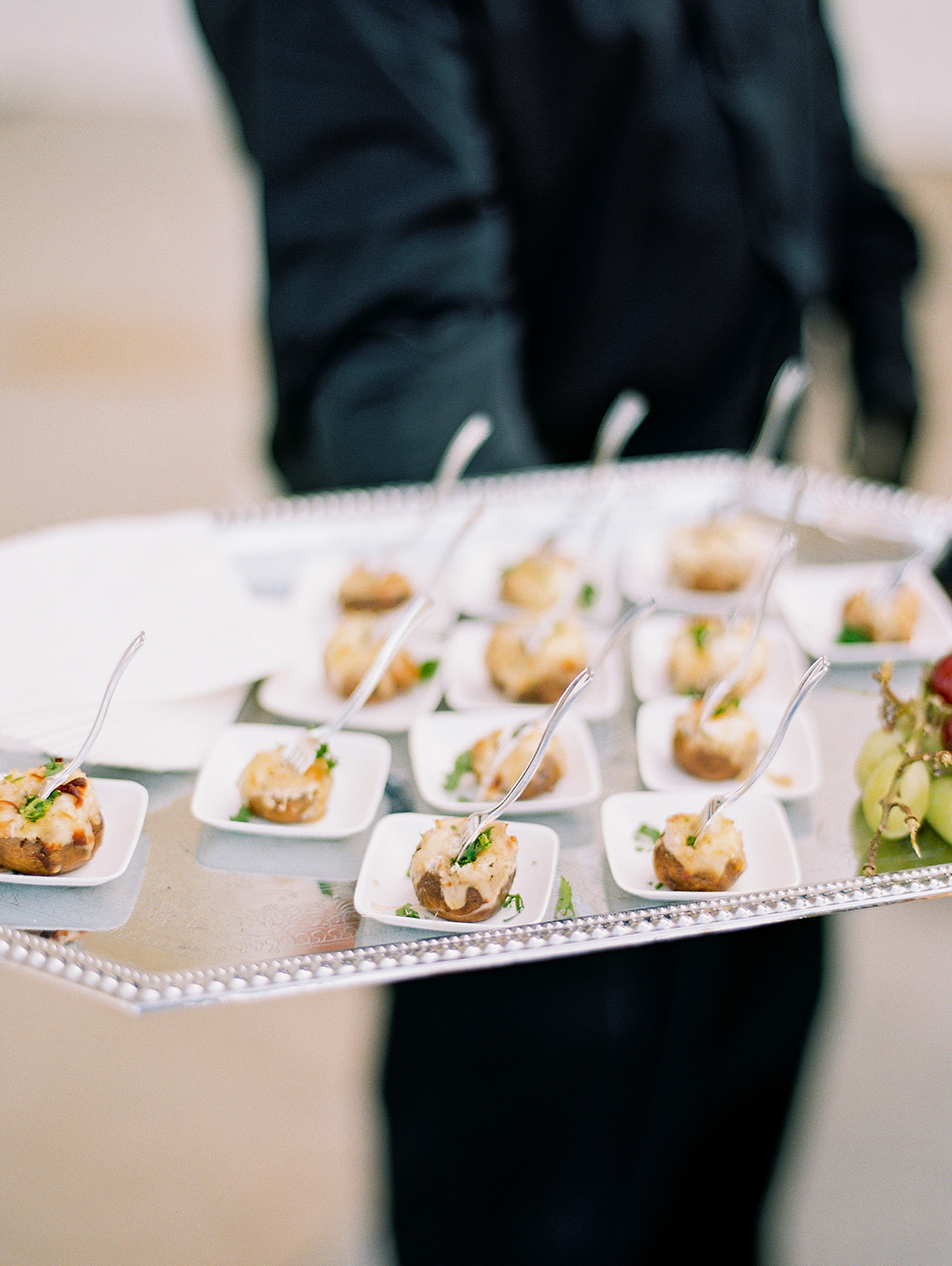 A server in black attire holds a silver tray with small white dishes, each containing an appetizer and a tiny fork. The tray also has a small bunch of grapes.