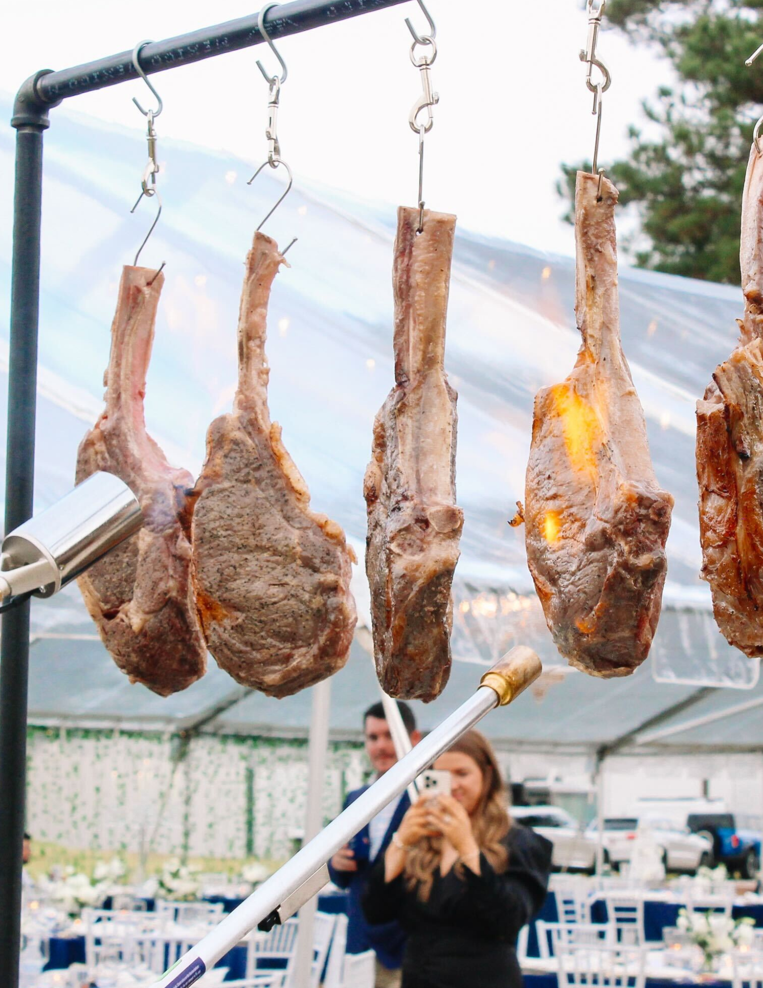 Fun Wedding Catering Five large tomahawk steaks hanging vertically on hooks, cooking over open flames, with an outdoor dining setup and two people in the background.