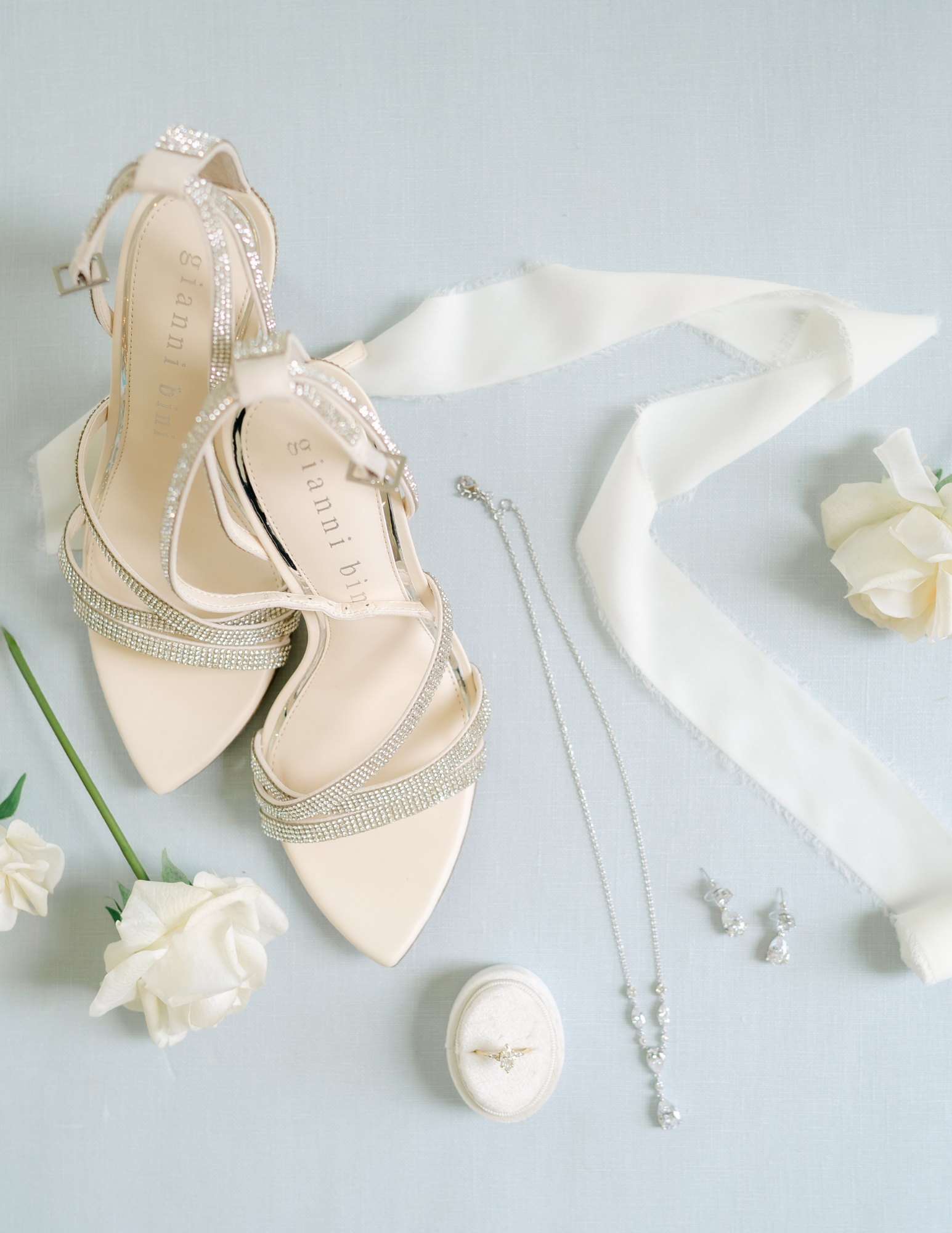 Arkansas Wedding Vendors Pair of elegant strappy heels, a necklace, earrings, a ring in a box, ivory ribbon, and white roses arranged on a light surface.