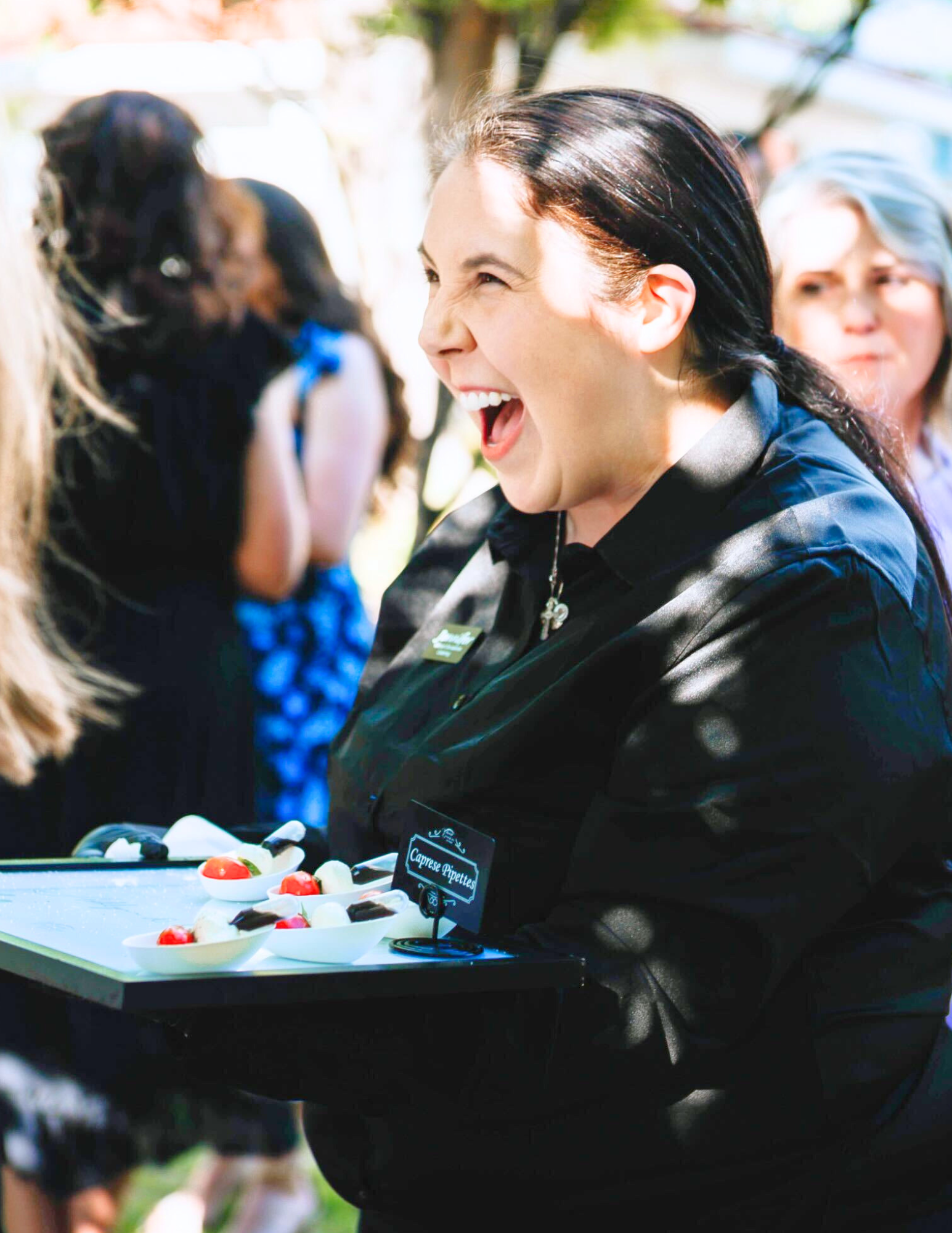 Arkansas Wedding Caterers A smiling server in black uniform holds a tray of hors doeuvres at an outdoor event, with guests and greenery blurred in the background.
