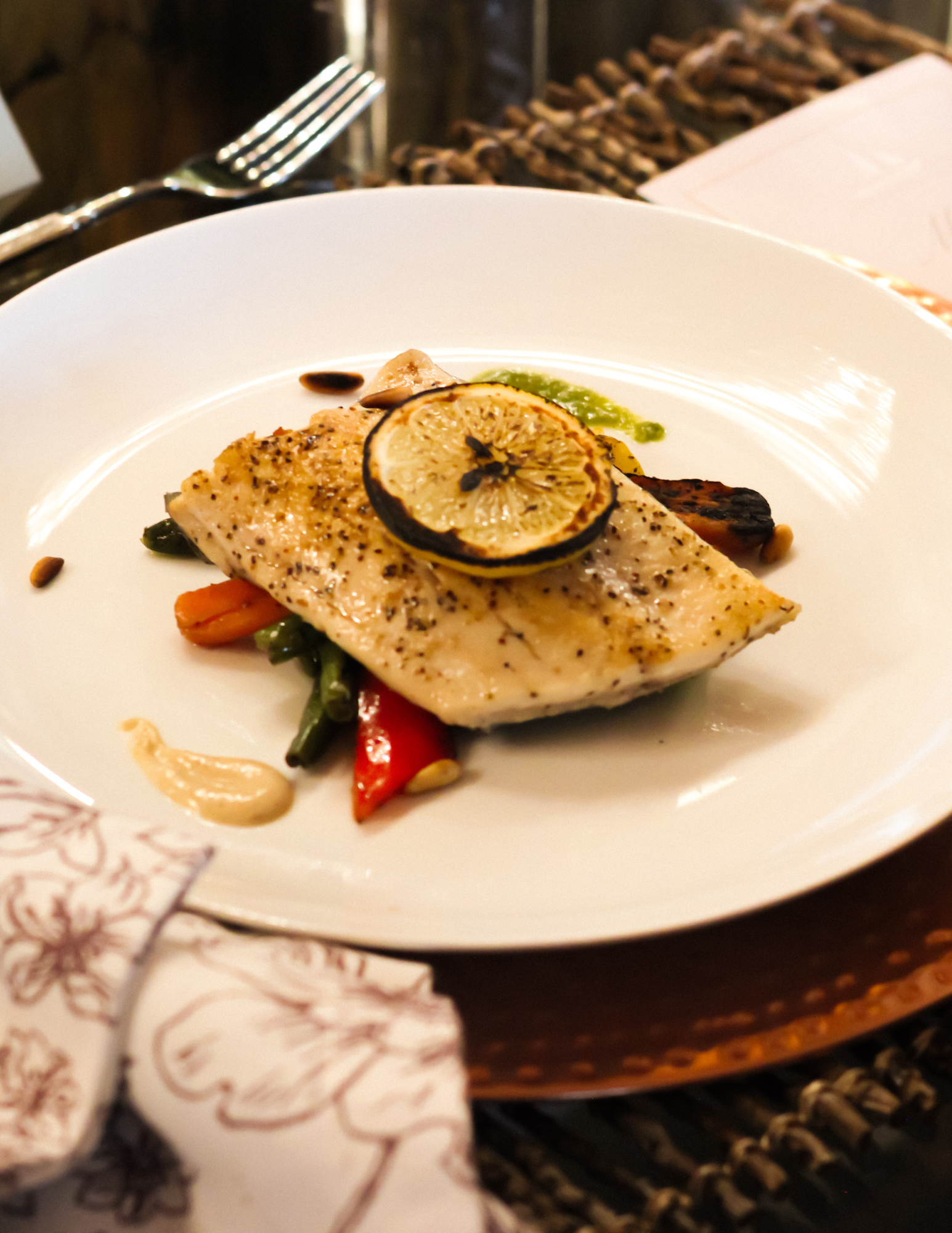 Party Catering Arkansas A plated dish featuring seasoned fish topped with a slice of grilled lemon, served over mixed vegetables and garnished with sauce. A napkin and fork are visible beside the plate.