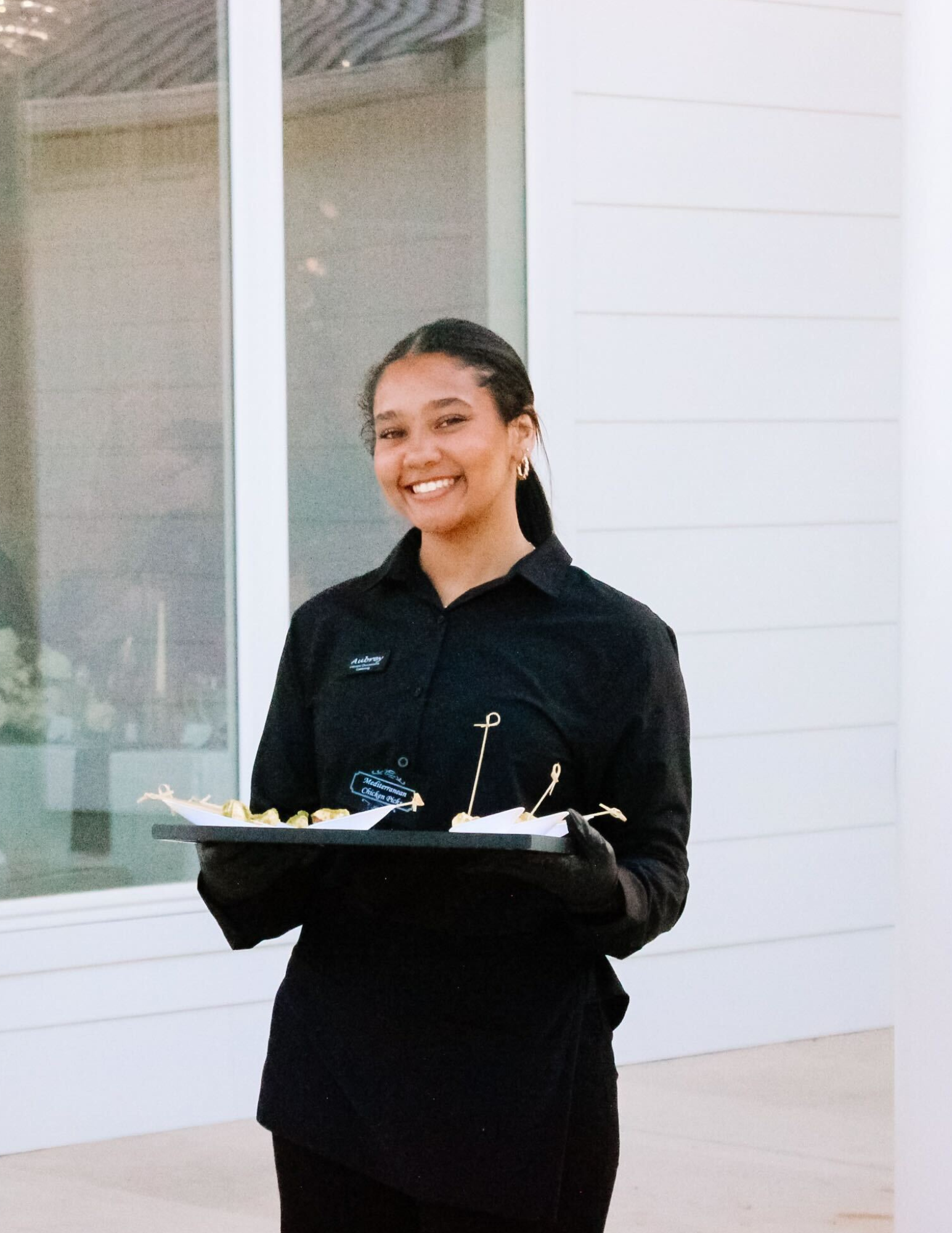 Passed Appetizers Catering A smiling server in a black uniform and gloves holds a tray of appetizers in front of a modern white building with large windows.
