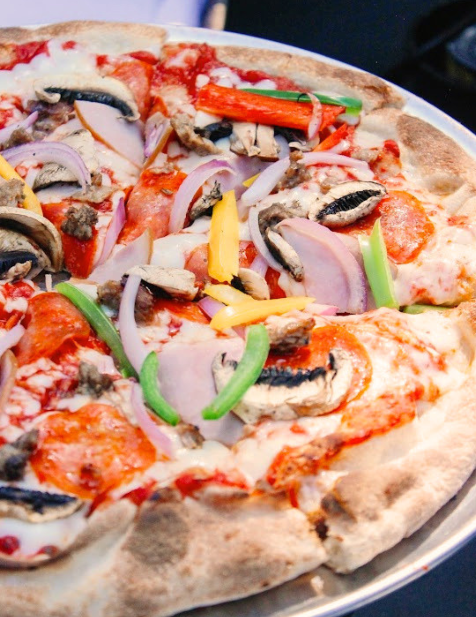 Social Party Catering Near Me A pizza with a thick crust, topped with pepperoni, sliced mushrooms, red onions, green and yellow bell peppers, and melted cheese, served on a metal tray.