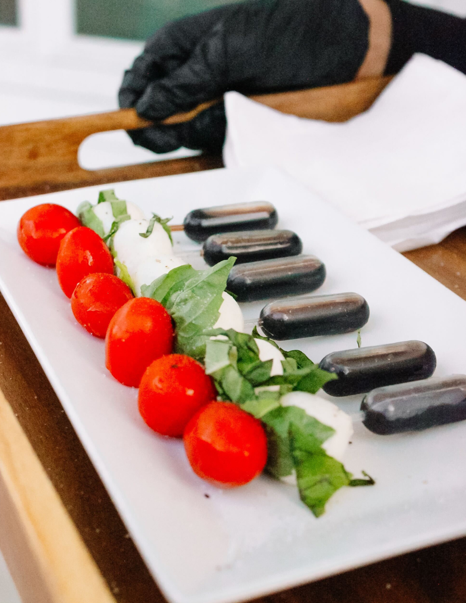 Appetizers Catering Arkansas A rectangular plate with cherry tomatoes, mozzarella slices, fresh basil leaves, and small dark sausages arranged in rows. A hand in a black glove holds a wooden tray nearby. White napkins are in the background.