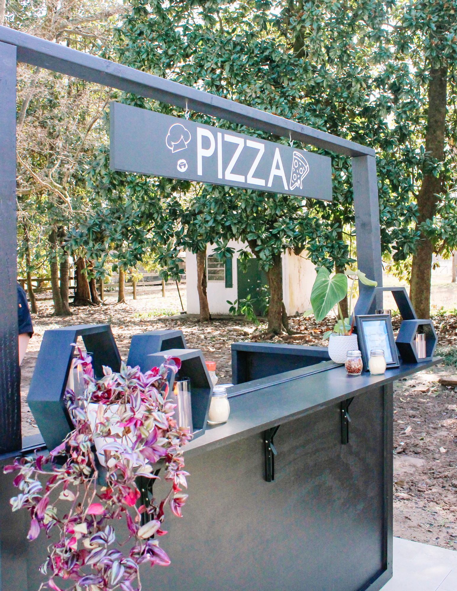 Pizza Social Catering Outdoor pizza stand with a black counter, geometric shelves, and a PIZZA sign above. There are potted plants and candles on the counter. Trees and a small white building are visible in the background.