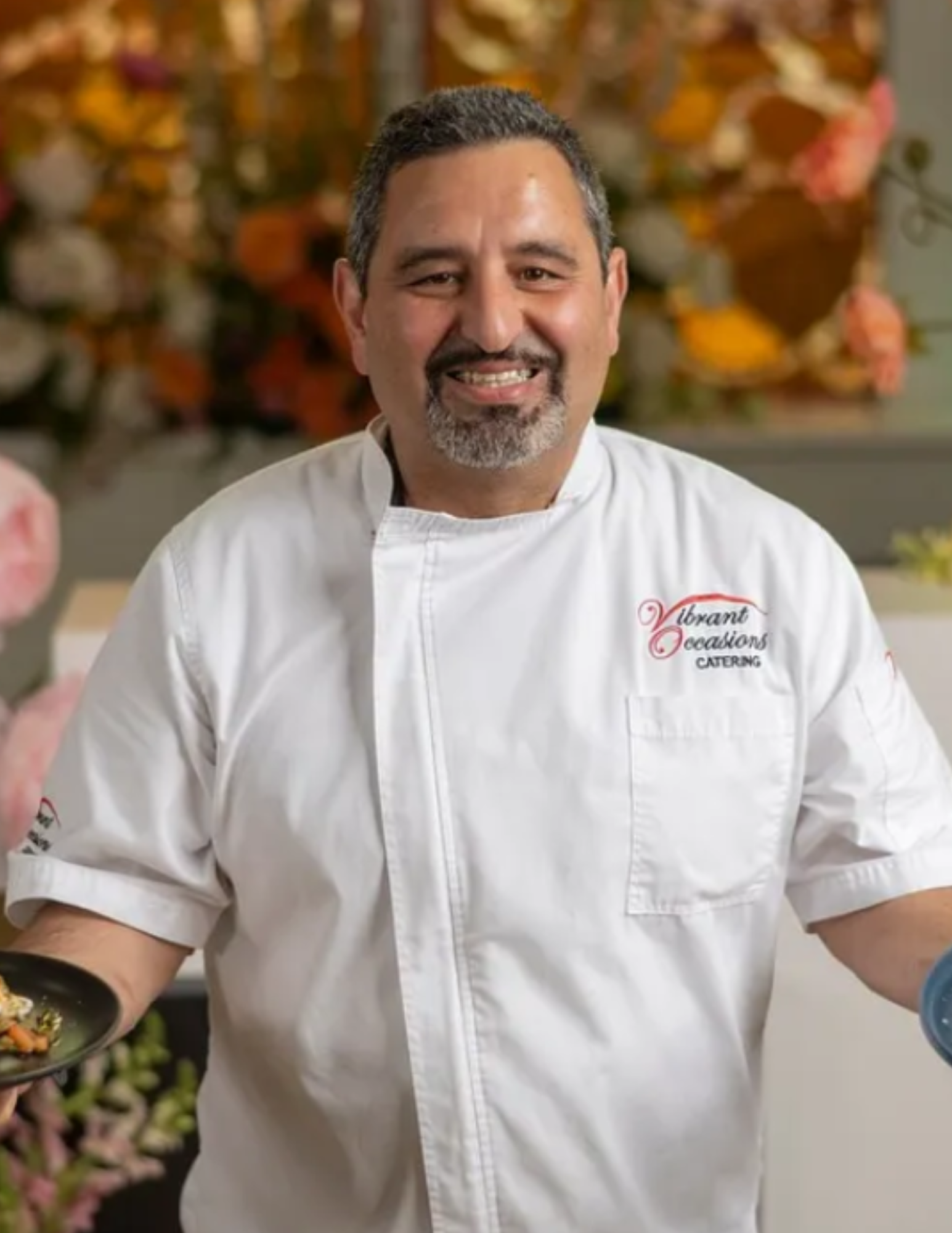Catering Socials A smiling chef in a white coat that reads Vibrant Occasions Catering stands indoors, holding two plates of food. There are blurred flowers and lights in the background.