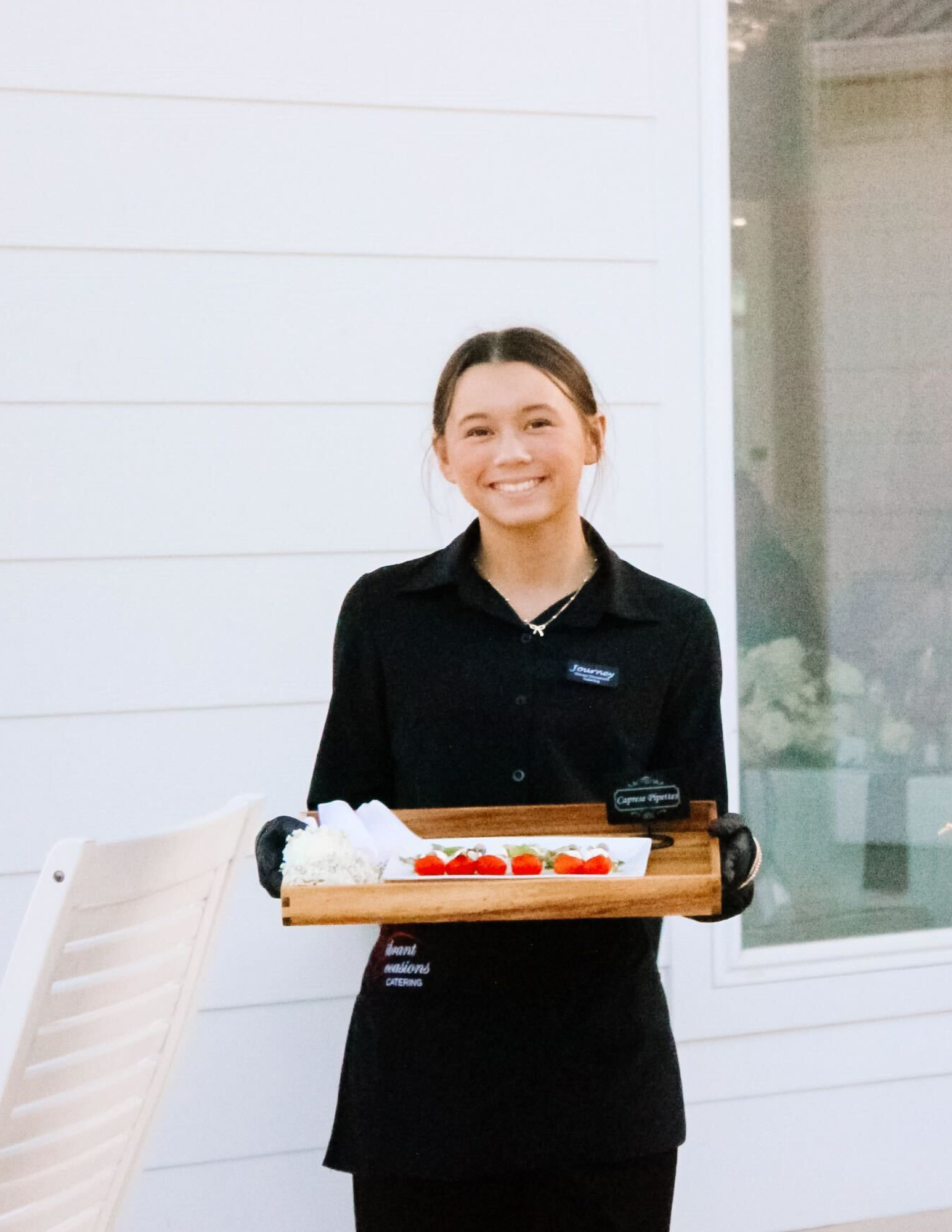 Business Catering Arkansas A smiling server in a black uniform holds a wooden tray with appetizers and napkins, standing outdoors by a white wall and chair.