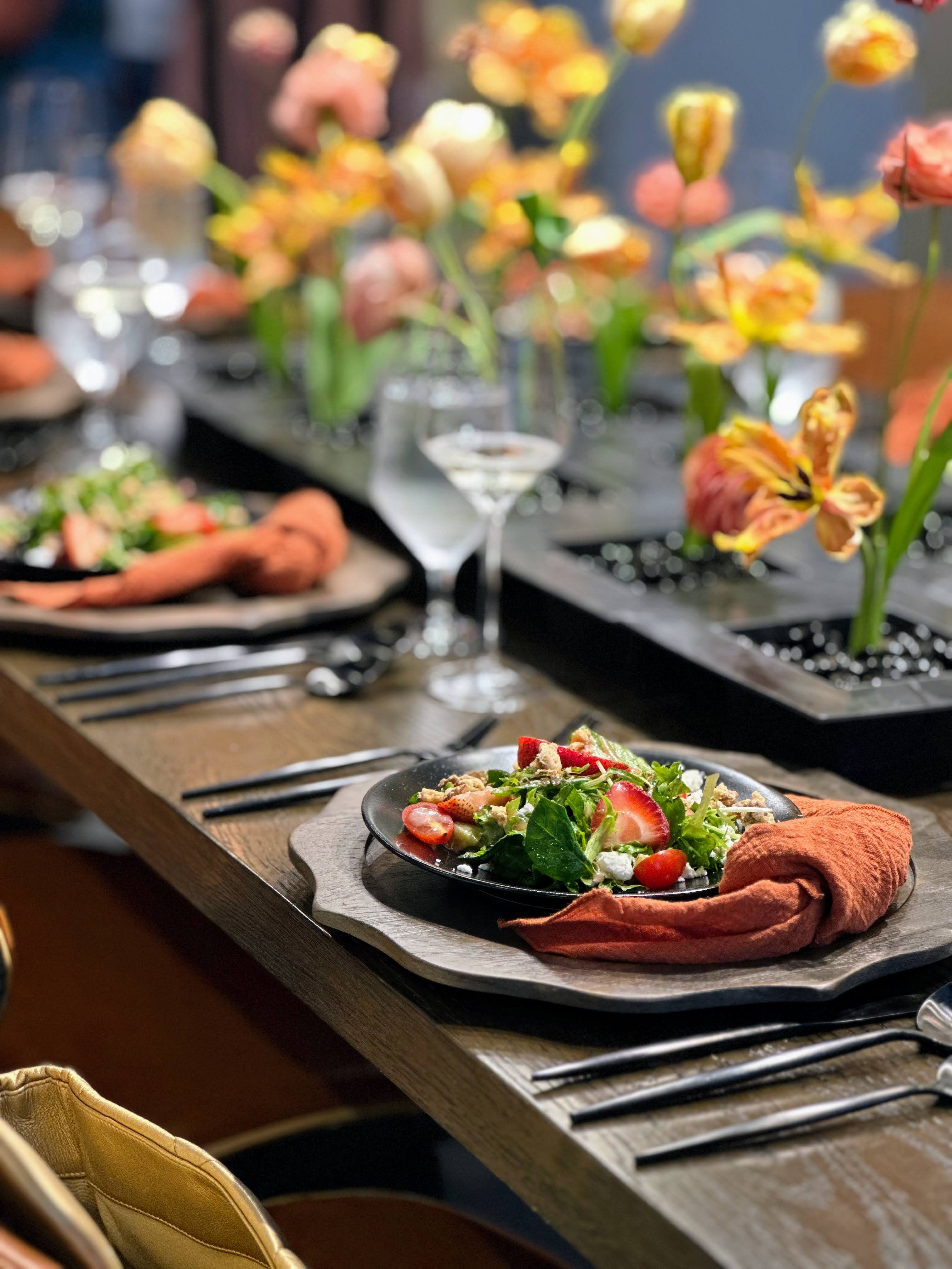 IMG_2027 A close-up of a beautifully set dining table with fresh salad plates, orange napkins, cutlery, water glasses, and a centerpiece of colorful flowers.