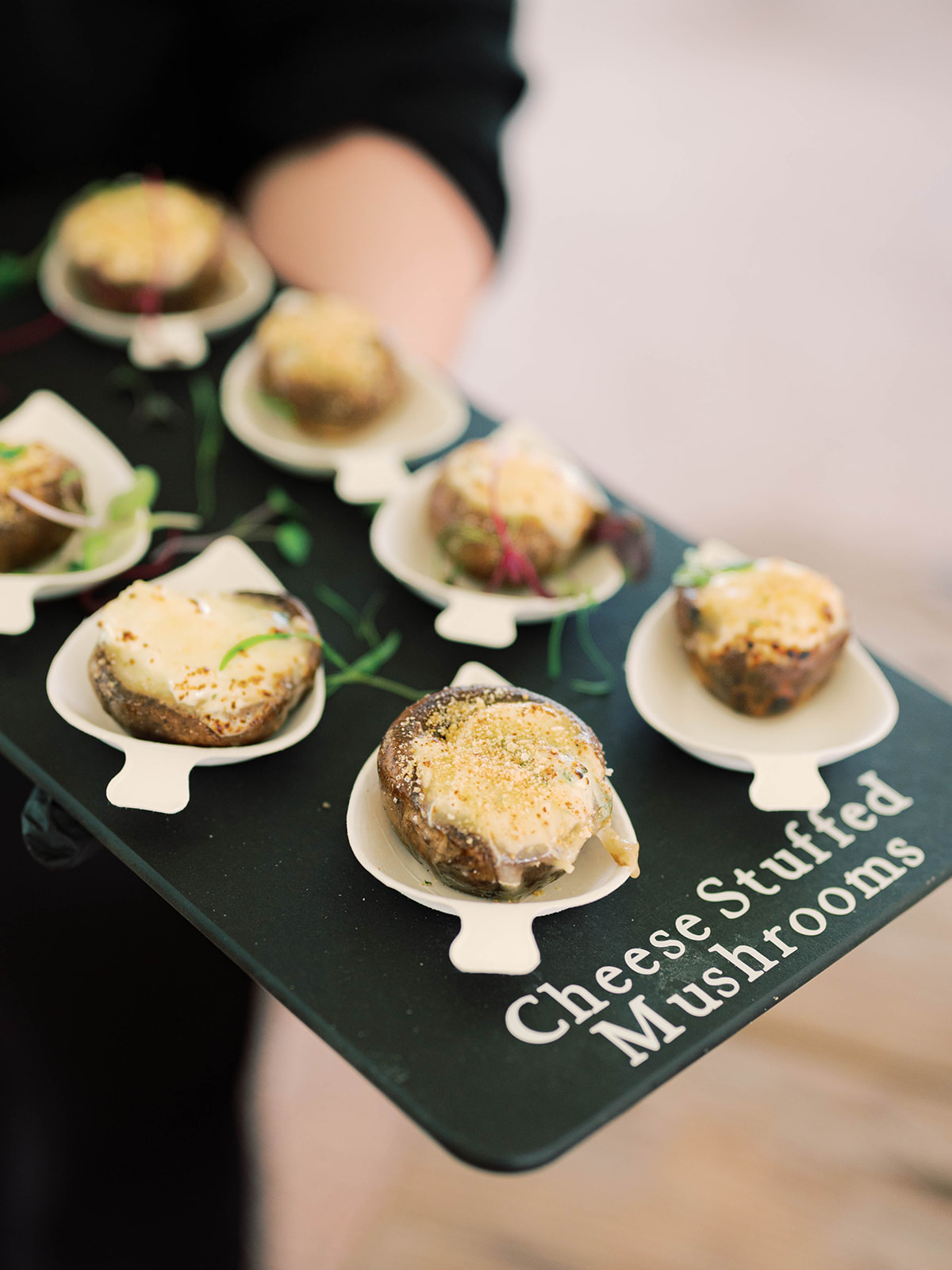 Liz and Matt Wedding A person holds a tray labeled Cheese Stuffed Mushrooms, featuring several mushrooms filled with melted cheese and garnished with herbs.