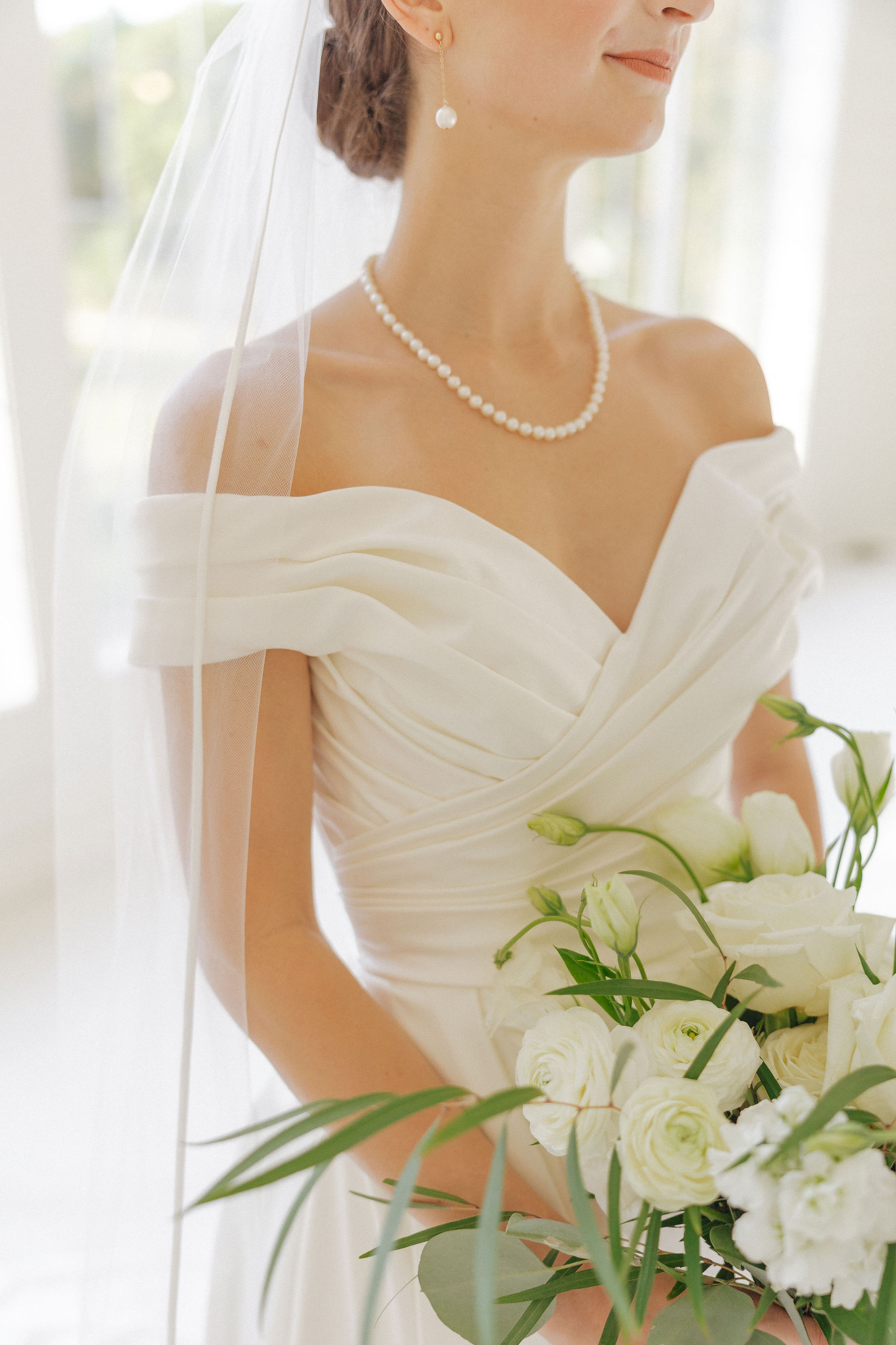 TaylorBridals-24745 Bride in an off-shoulder white gown with a pearl necklace and veil, holding a bouquet of white flowers and greenery, standing in bright, natural light.