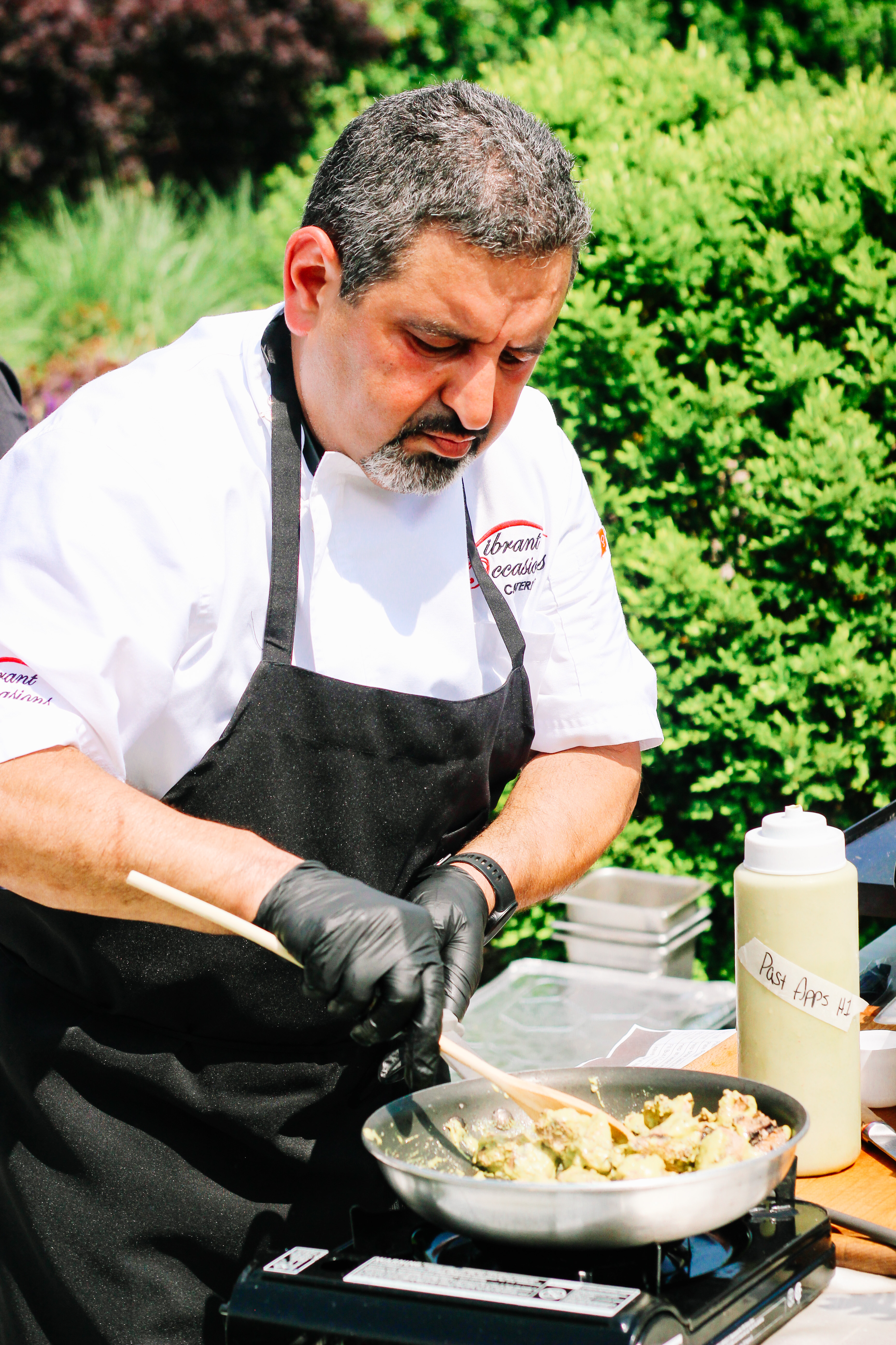 A chef wearing a white coat and black apron cooks food in a pan outdoors, stirring with a wooden spoon. Bottles and greenery are visible in the background.