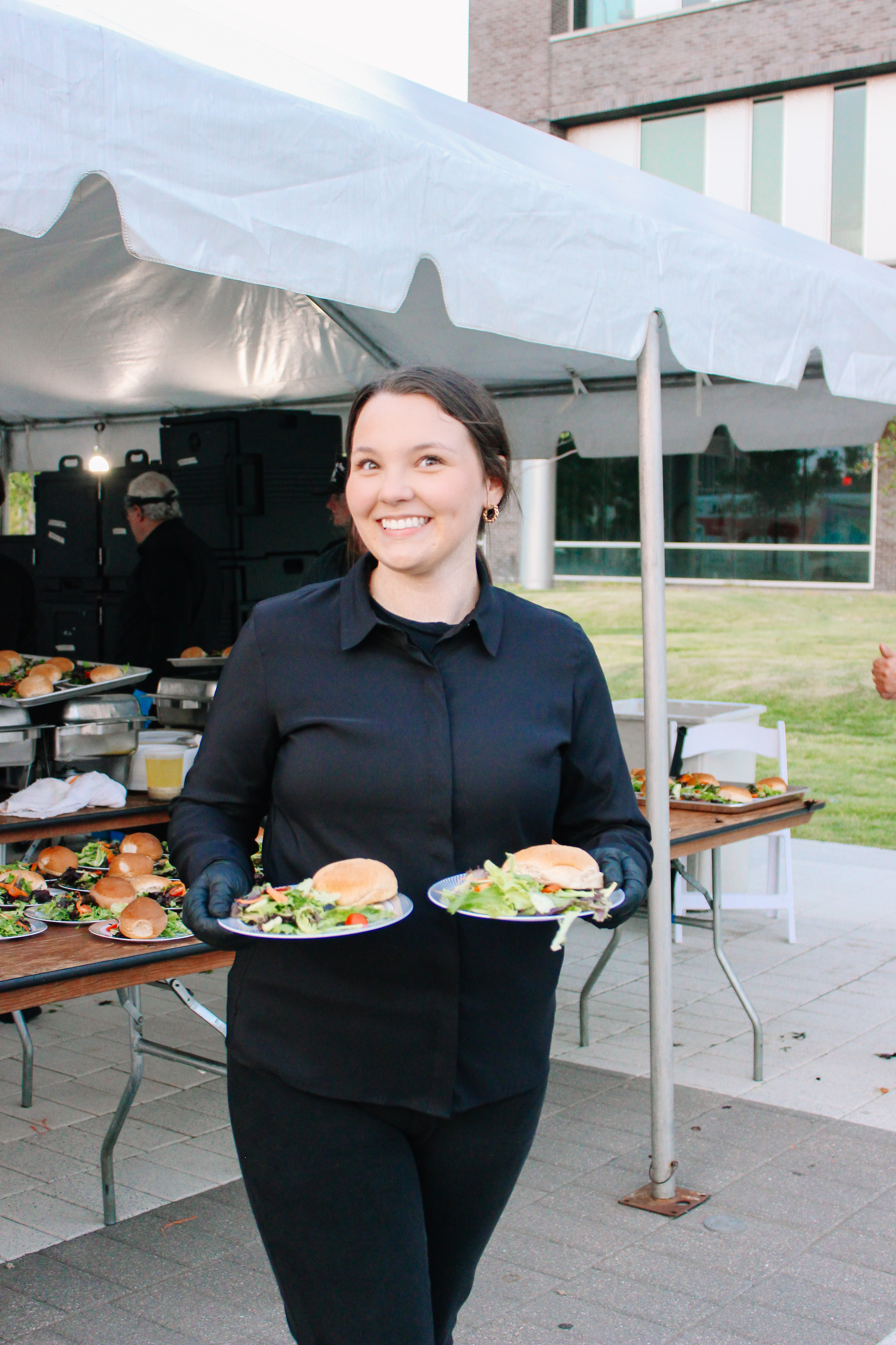 IMG_9625 A woman in black clothing smiles while holding two plates of food, each with a sandwich and salad, at an outdoor event under a white tent with tables of food behind her.
