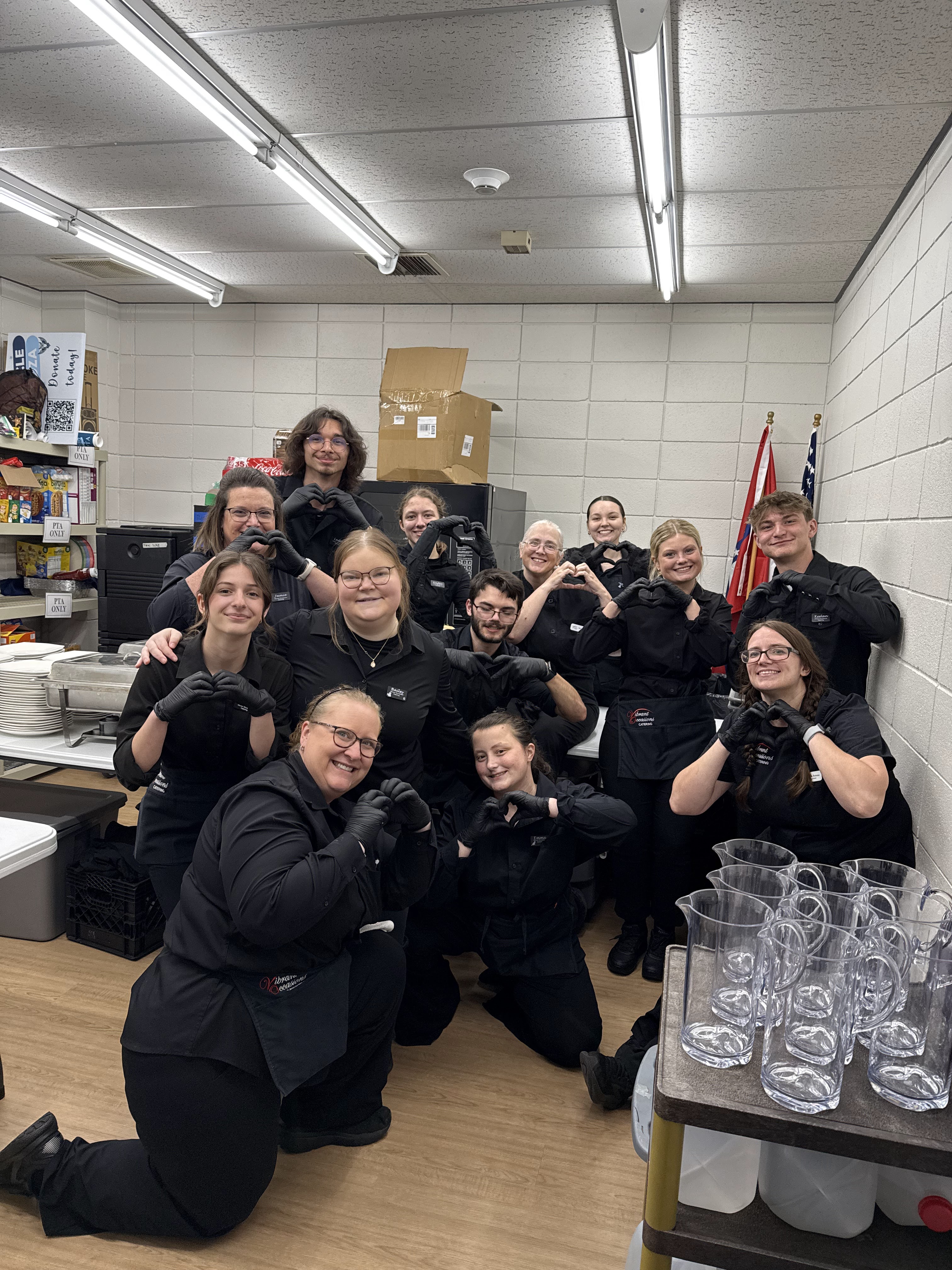 IMG_9740 A group of people in black uniforms pose and smile in a break room, making heart shapes with their hands. There are pitchers and supplies on a cart and shelves, with flags displayed in the background.
