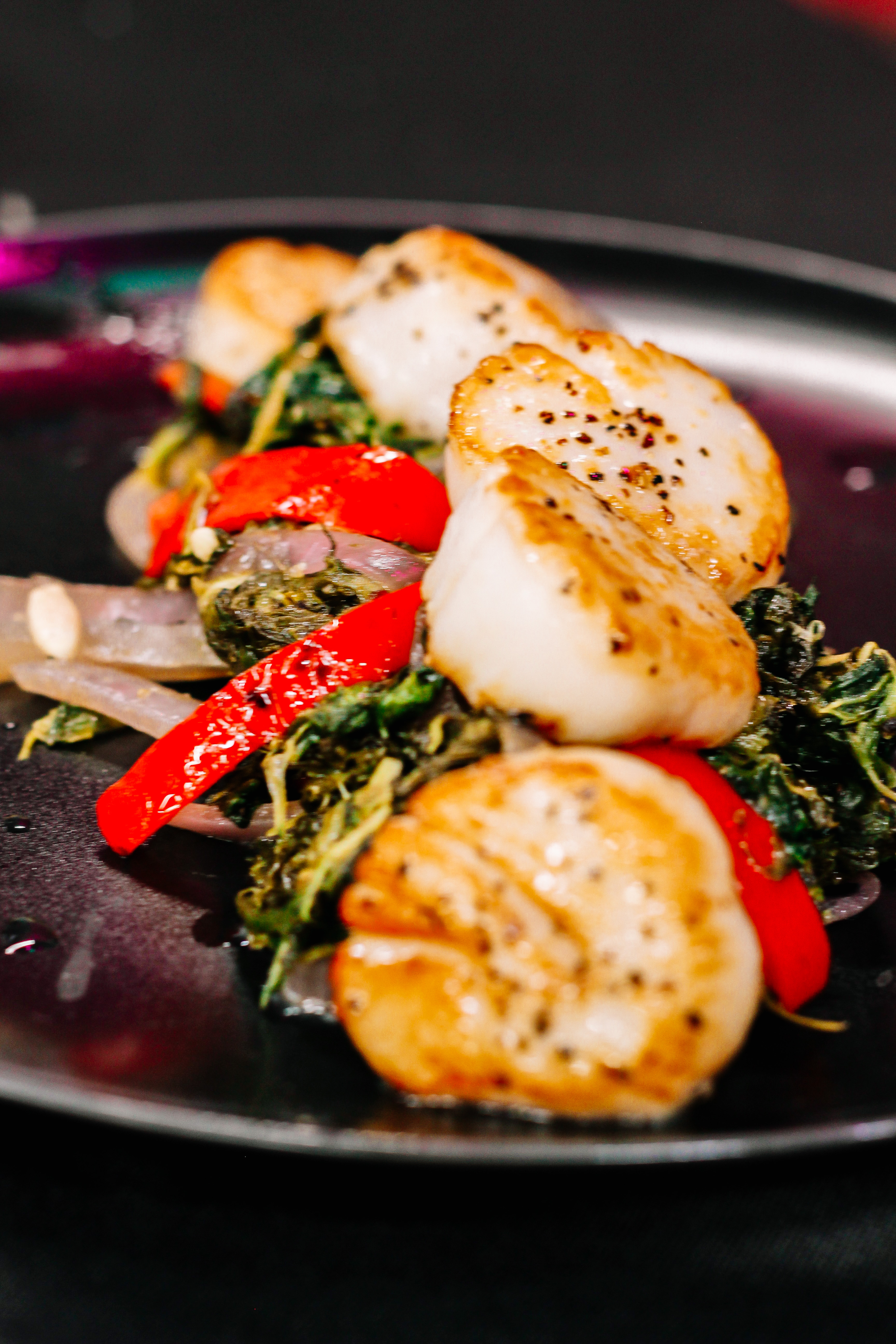 Pan Seared Scallops(2) (1) Seared scallops served on a bed of sautéed greens, red bell peppers, and onions, presented on a dark plate.