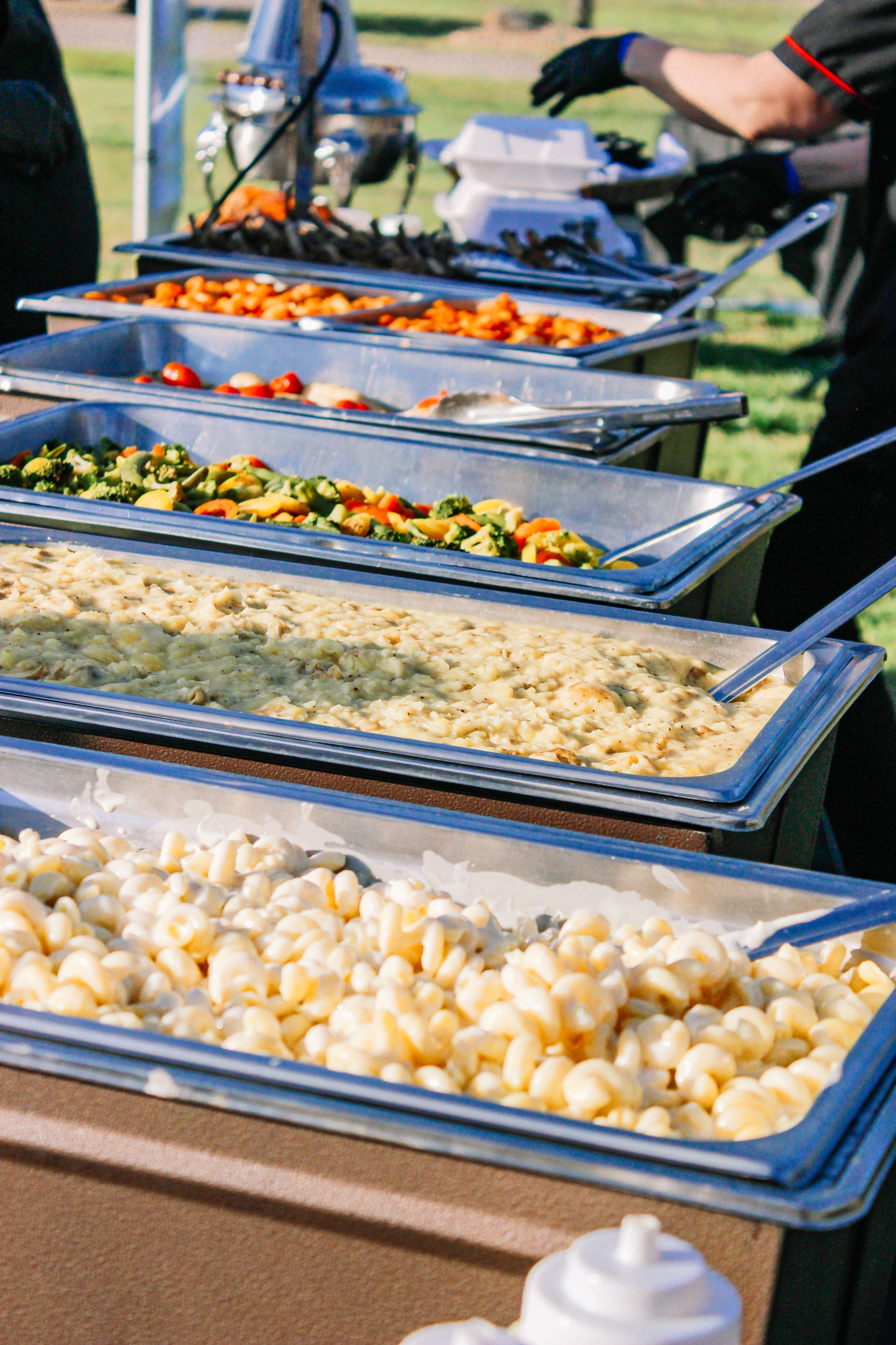 A buffet table outdoors displays trays of gnocchi, pasta, vegetables, and various hot dishes. A person in black gloves serves food in the background.