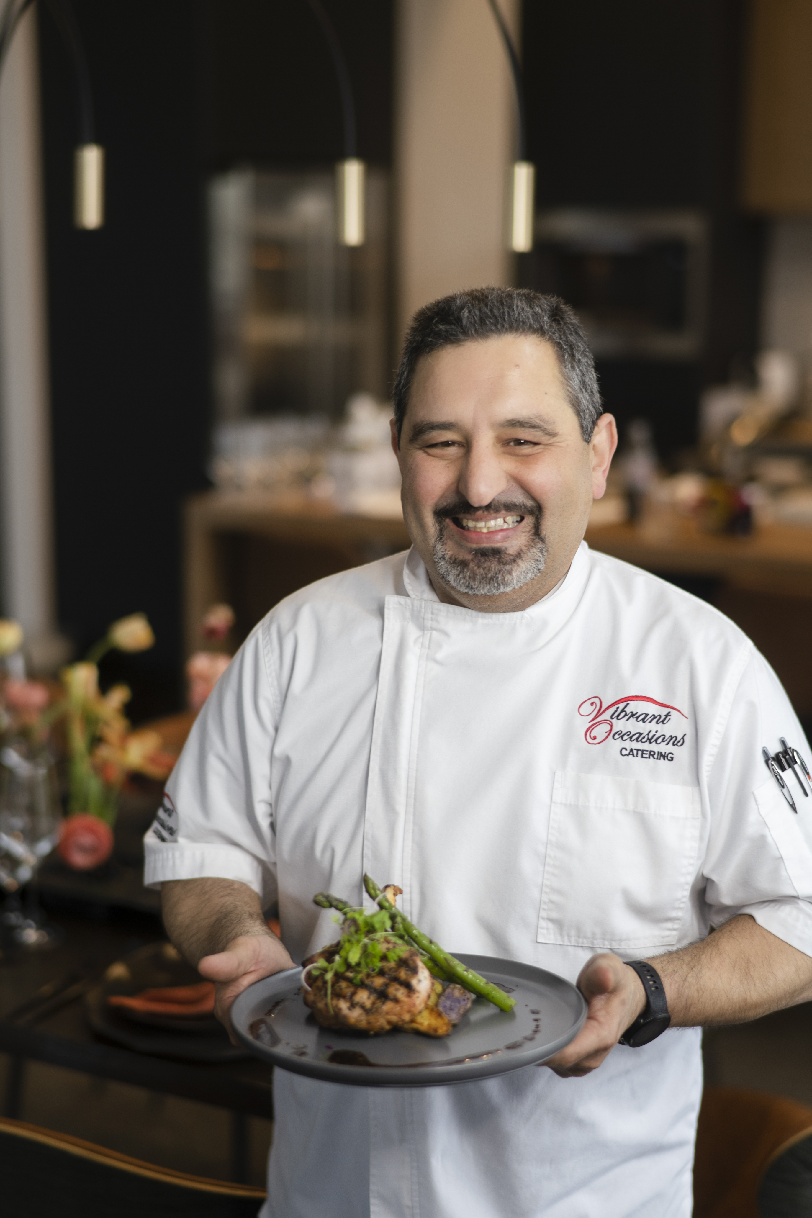 Chef Serge Krikorian A smiling chef in a white coat holds a plated dish topped with asparagus. The background shows a blurred dining area with elegant table settings.