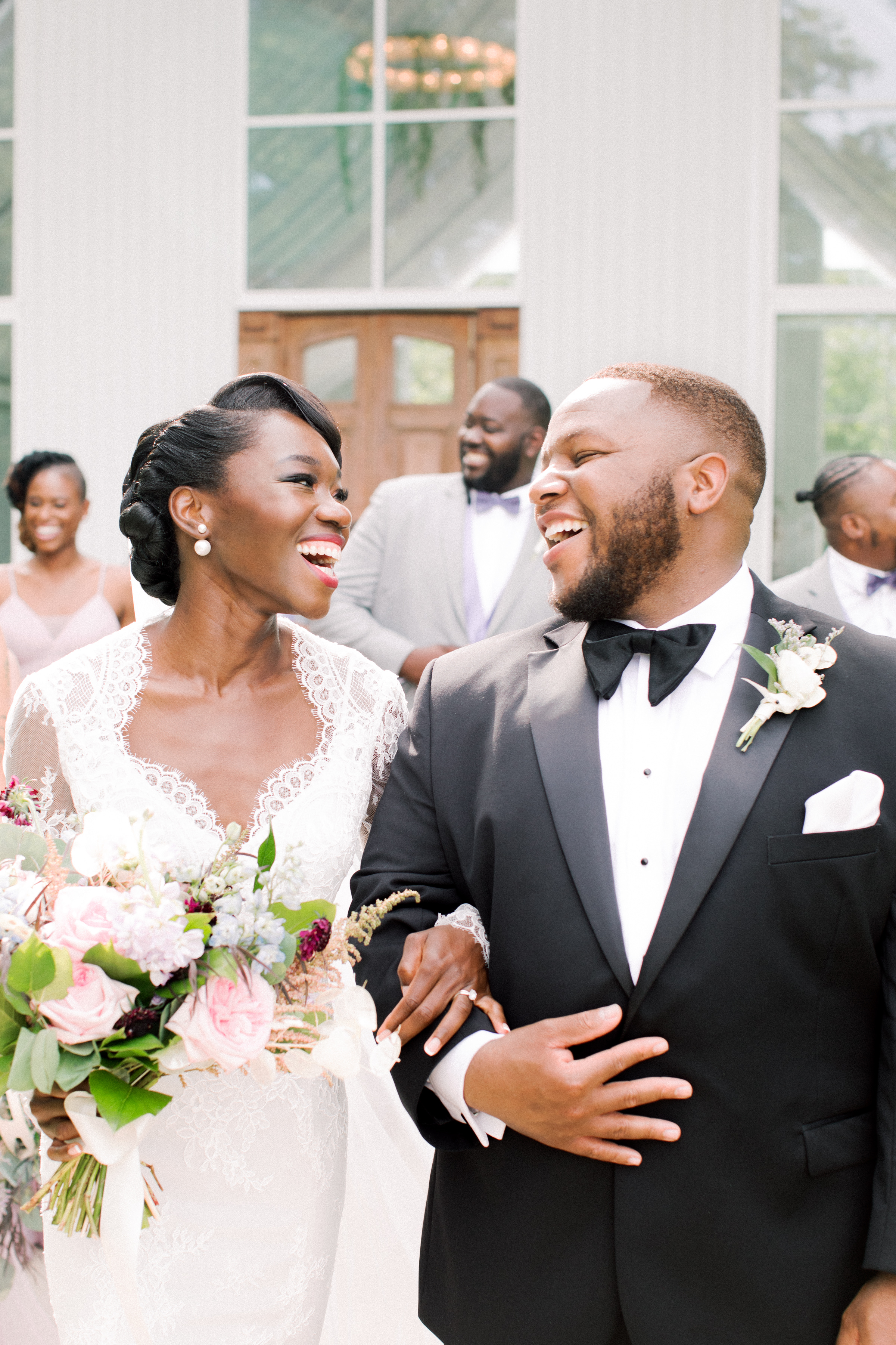 Rebecca + Kevin A bride and groom smile and laugh as they walk arm in arm outside, surrounded by their joyful wedding party. The bride holds a bouquet of flowers and wears a lace dress; the groom is in a black tuxedo.