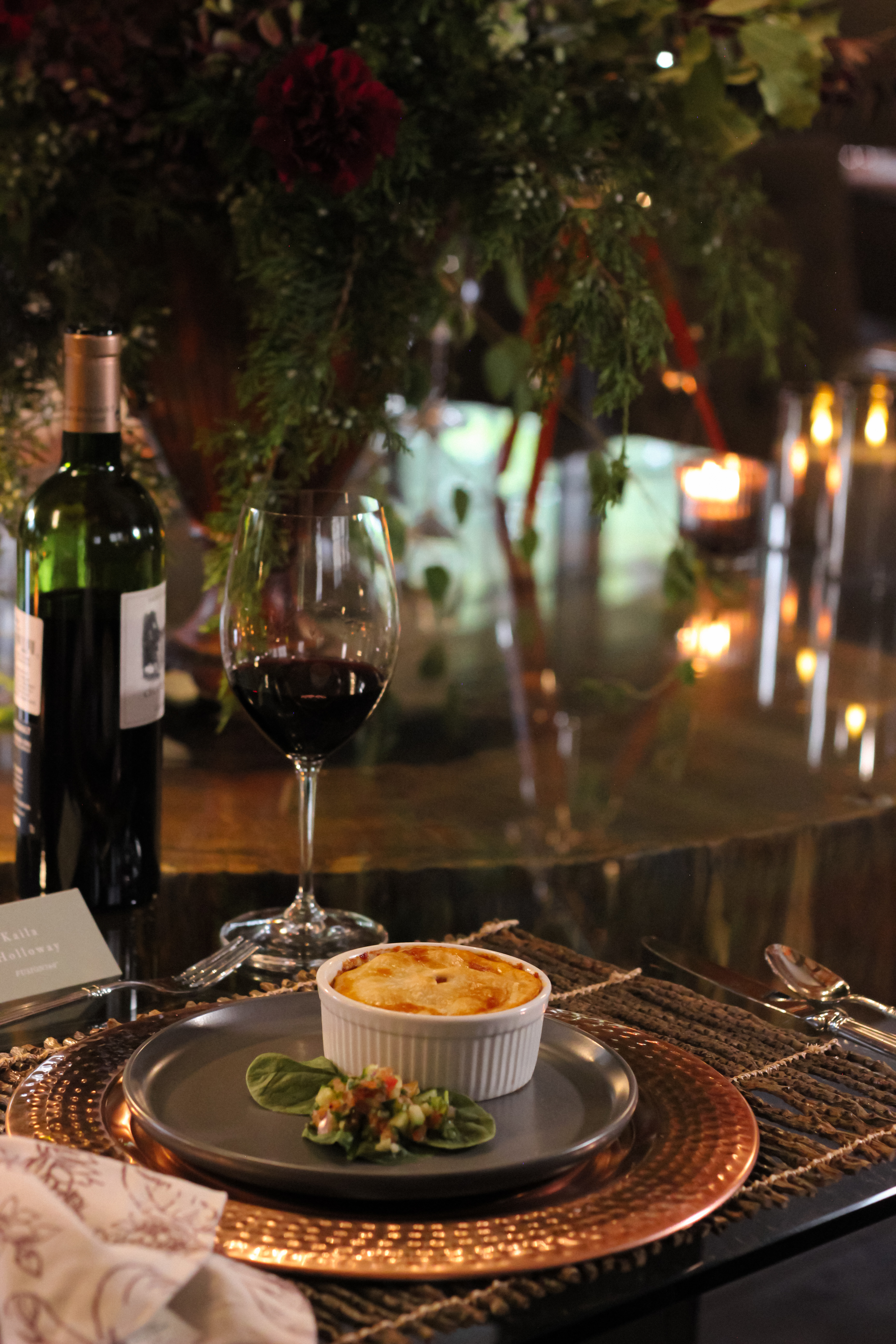 A ramekin with a baked dish and a small side salad on a dinner plate, accompanied by a glass of red wine and a bottle of wine.