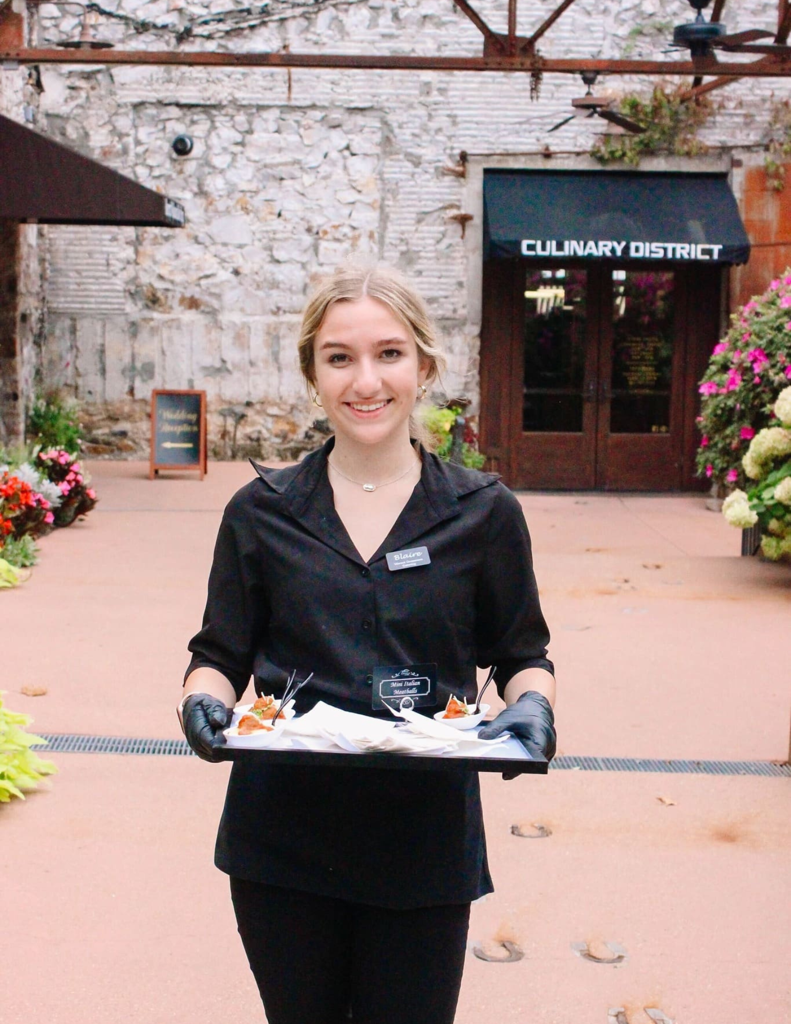 Arkansas Catering Staff A smiling server in a black uniform and gloves holds a tray of appetizers in front of a building entrance labeled Culinary District.