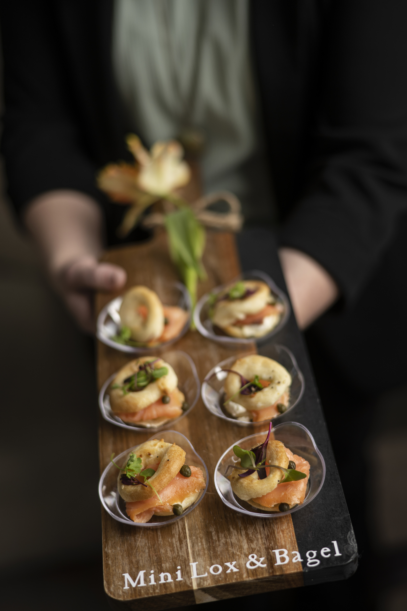 A wooden serving board held by a person, featuring several mini lox and bagel appetizers garnished with capers and greens, with text on the board reading Mini Lox & Bagel.